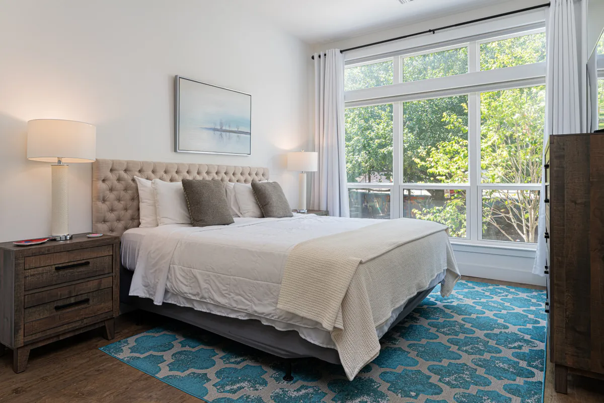 This bedroom scene displays a plush bed with large windows that let in natural light, elegant nightstands, a decorative rug, and artwork on the walls. The overall mood is tranquil and inviting, hinting at restful moments after a productive day.