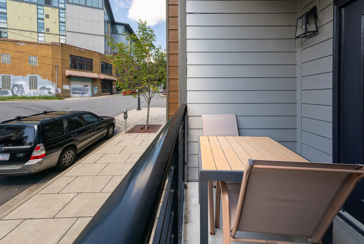 The image shows a cozy outdoor table and chairs overlooking a street view, with the building's facade in the background. The setting is relaxed, perfect for sipping coffee, reading a book, or socializing while taking in the energy of the city.