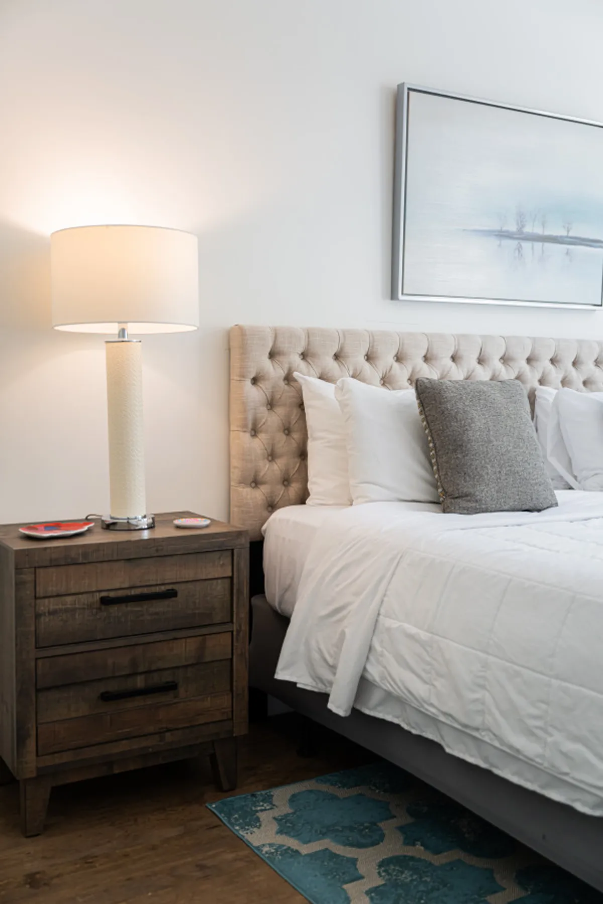 A serene king bedroom featuring a bed with a tufted headboard, plush linens in soothing tones, a nightstand with a lamp for gentle lighting, and decorative artwork above the bed. The soft bedding and calming color palette create a sense of tranquility and comfort.
