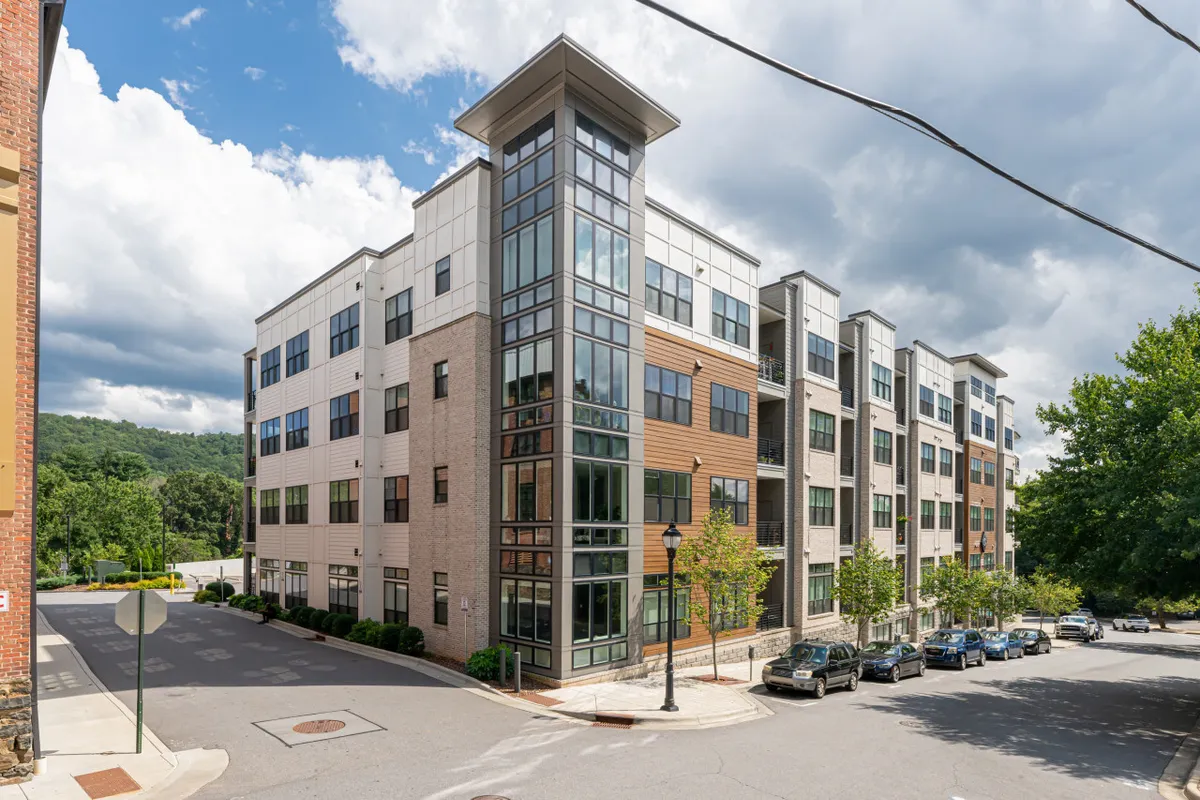 A striking exterior view of a modern residential building with large glass windows and landscaped greenery surrounding the entrance. The architecture is contemporary and inviting, with hints of communal activity and easy access to the outdoors.