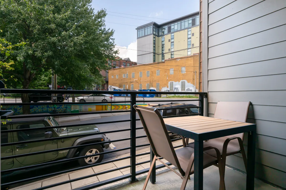 A private balcony furnished with a small table and chairs, overlooking a lively street lined with trees. The space is casual and inviting—perfect for sipping coffee, reading, or enjoying the city’s vibrant atmosphere from an urban oasis.