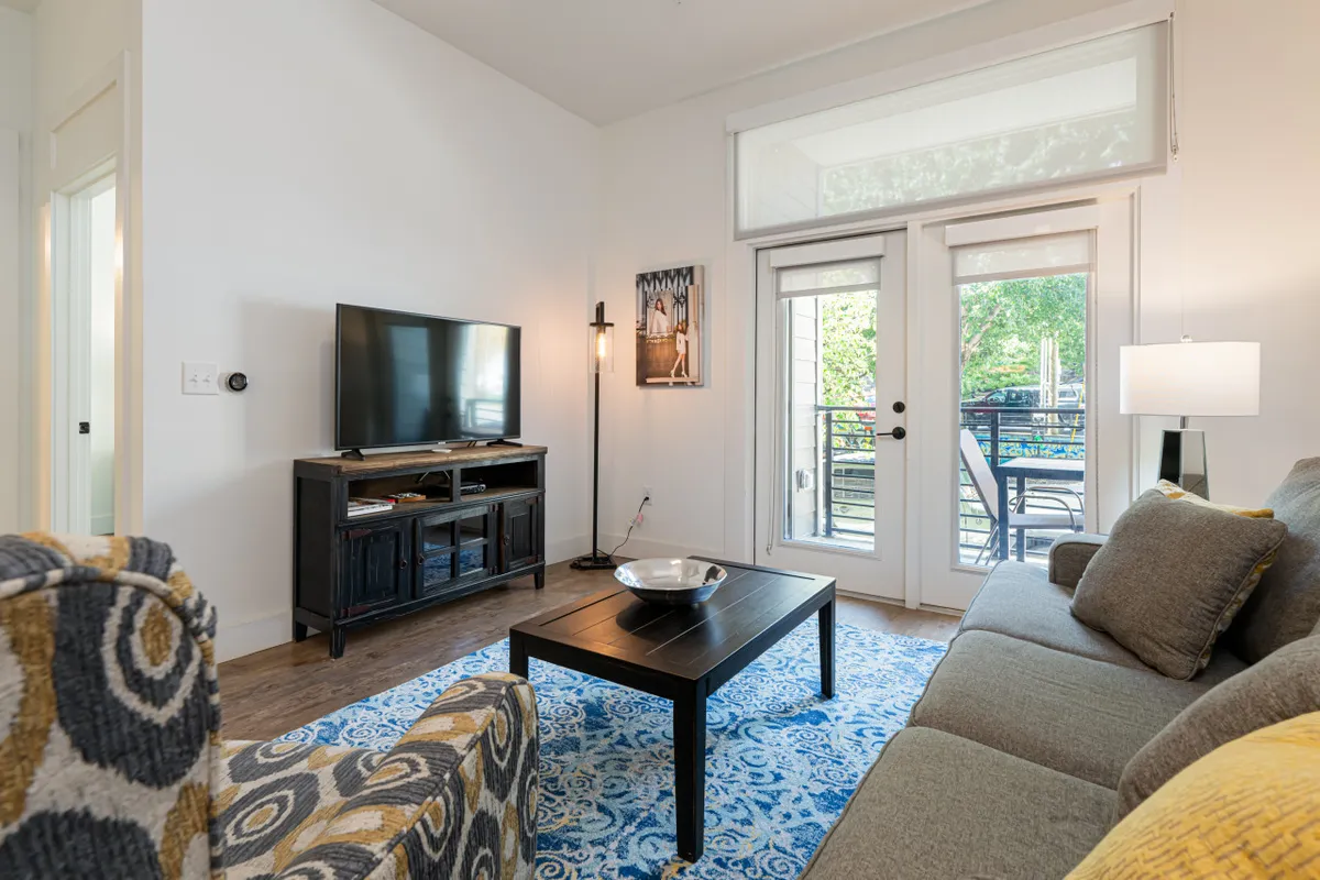 The image showcases a bright, inviting living area with a plush sofa facing a large television, a stylish coffee table, and expansive windows that fill the space with natural light and offer views of the outdoors. The open layout hints at a seamless transition to the balcony, and the ambiance is one of comfort and relaxation—perfect for enjoying a cozy movie night or basking in the morning sun.