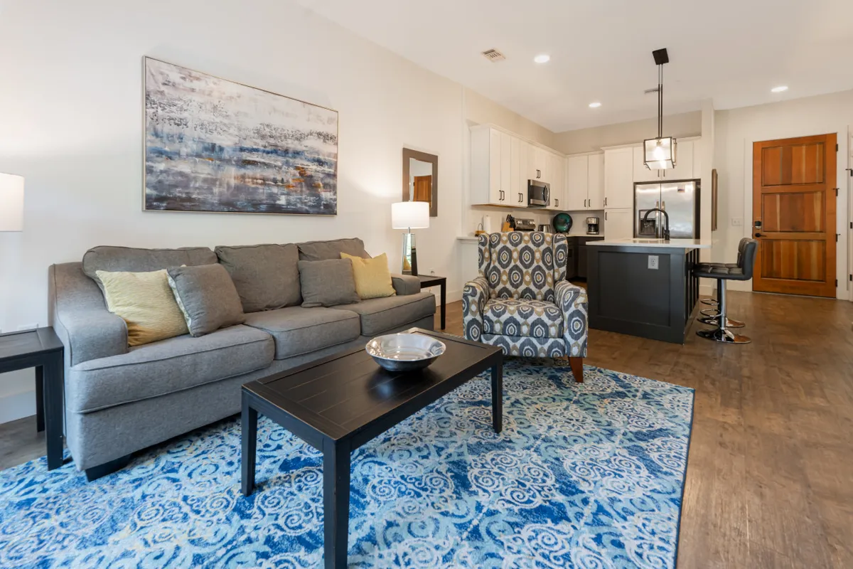 A modern, open-plan layout showing a gray sofa, colorful area rug, accent chair, and a glimpse of the kitchen island in the background. The space is cohesively arranged for both relaxation and entertaining, emphasizing the seamless flow between living and dining areas.