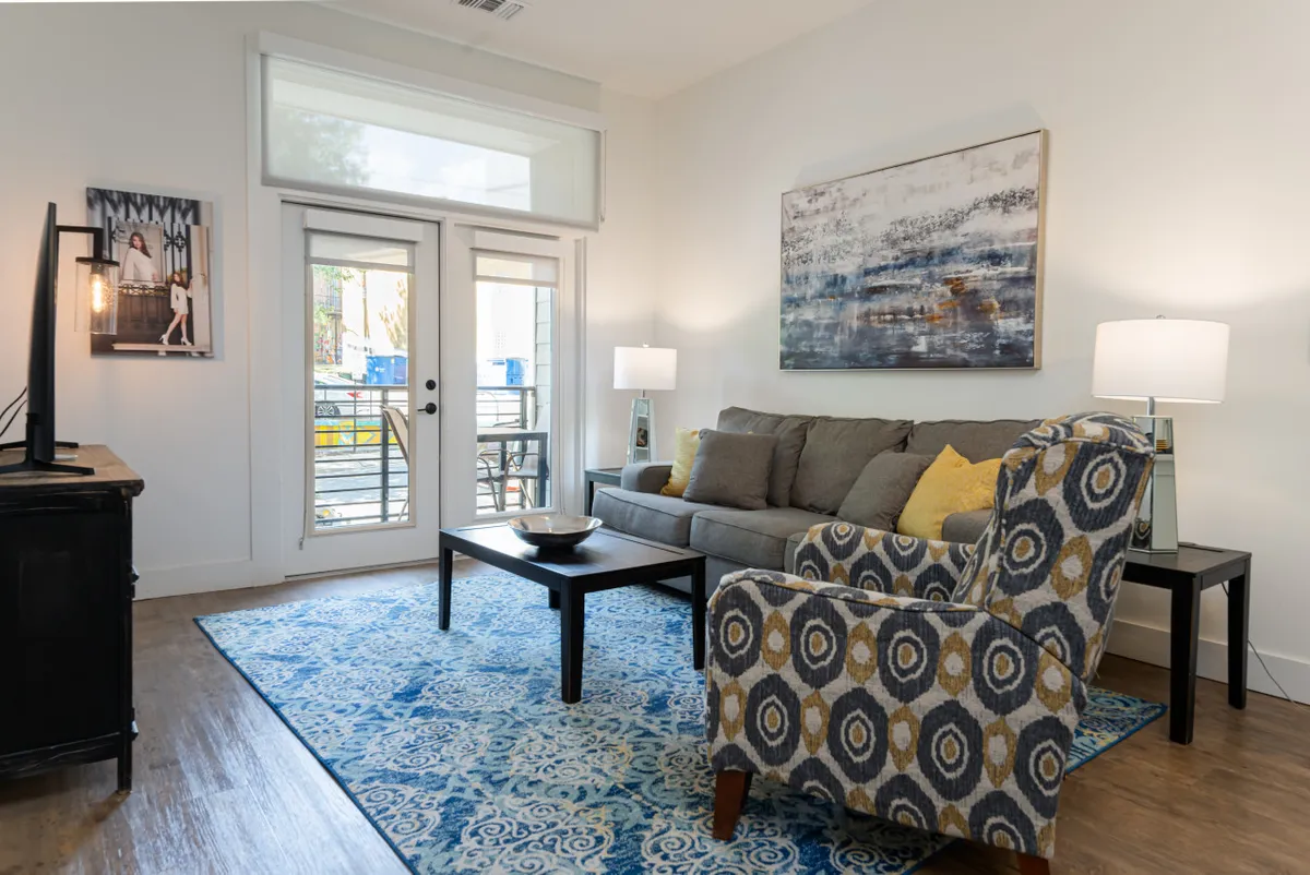 A bright, comfortable living room featuring a plush sofa, accent chair, central coffee table, vibrant artwork on the wall, and large windows that flood the space with natural light. The arrangement is open and inviting, ideal for socializing or unwinding after a day out.