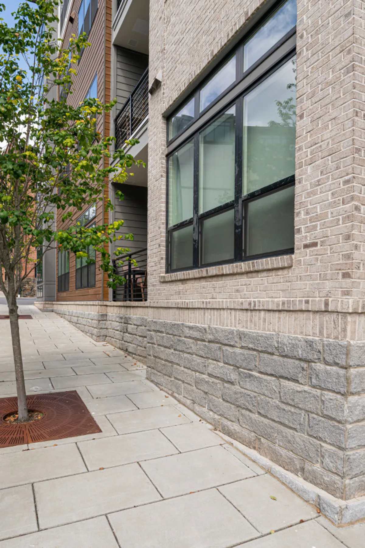 This image features the exterior of a modern building with large windows, a tree, and a brick and stone facade. People are seen strolling along the sidewalk and enjoying the vibrant outdoor environment, set against the backdrop of welcoming architecture.