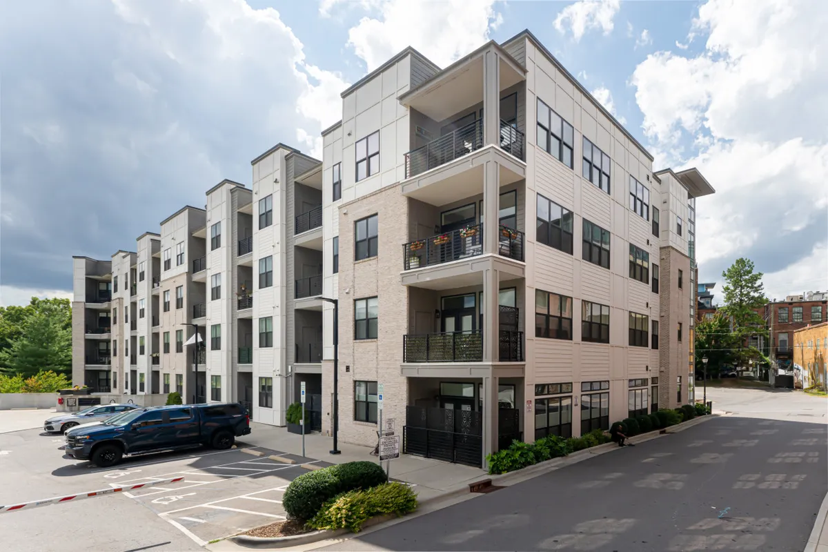 The photo captures the building's modern architecture, balconies with greenery, and a convenient, clearly visible parking area at ground level. The environment appears well-maintained and accessible.